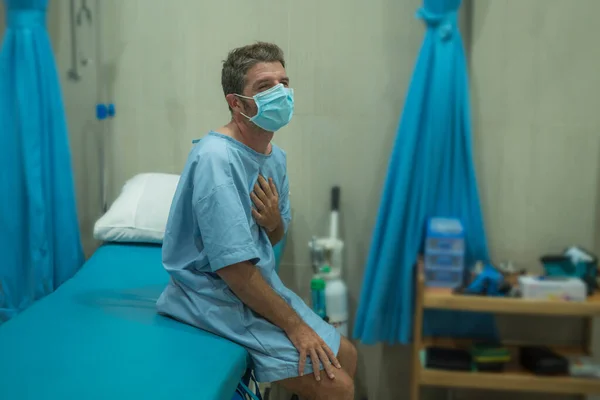 attractive and scared man infected by covid-19 - dramatic portrait of adult male in face mask receiving treatment at hospital suffering respiratory disease sitting on bed worried