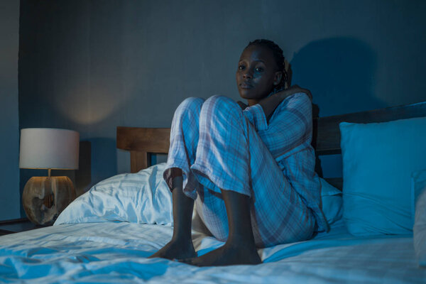 молодая грустная и депрессивная чернокожая афро-американка, сидящая дома на кровати несчастная и бессонная по ночам, чувствующая себя подавленной, страдая депрессией и бессонницей
