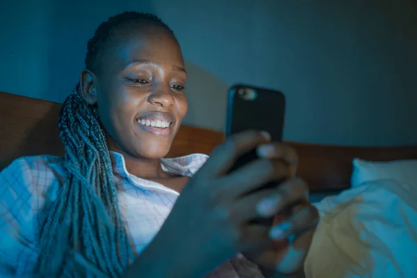 happy African American millennial woman as social media addict - night lifestyle portrait of young beautiful and cool black girl texting or online dating on mobile phone in bed smiling cheerful