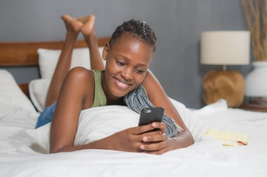 Genç, mutlu ve çekici zenci Afrikalı Amerikalı kadının portresi sosyal medya uygulamasını cep telefonunda rahat ve neşeli internet buluşmalarında kullanarak yatakta yatıyor.