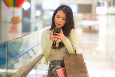 Modern alışveriş merkezinden alışveriş yapan genç ve güzel Asyalı bir kadın. Elinde alışveriş çantaları tutan mutlu ve çekici bir Çinli kız. Cep telefonuyla güzellik mağazasından alışveriş yapıyor.