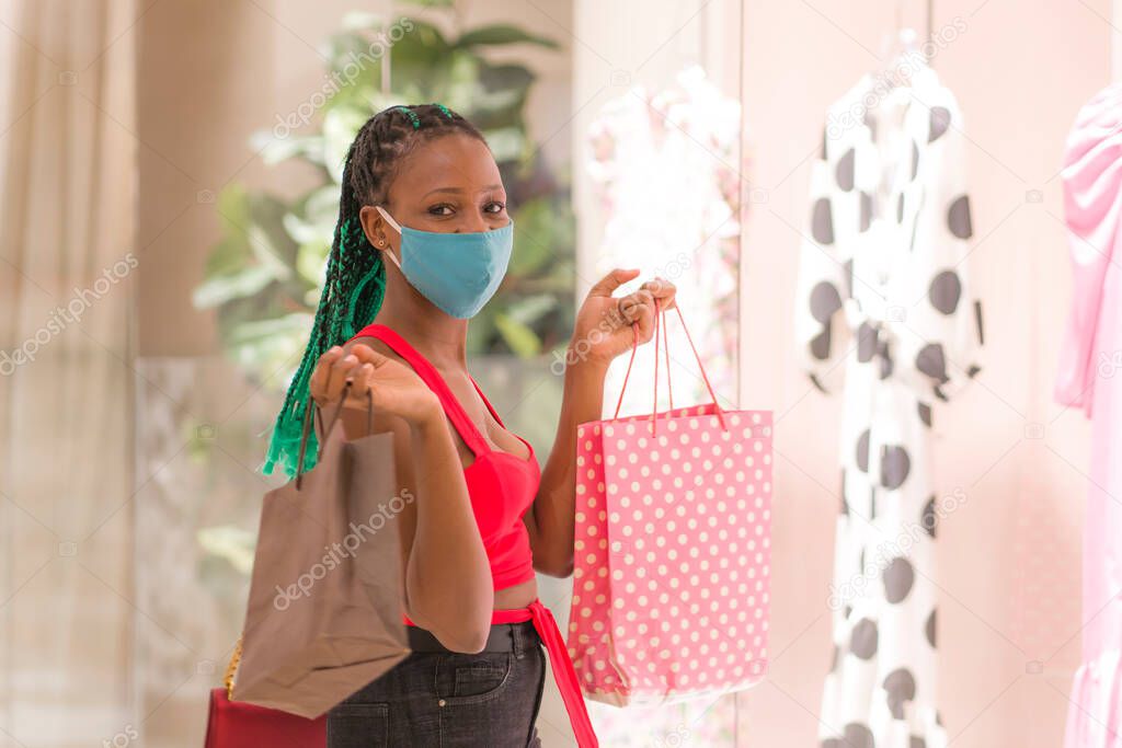 young African American woman at shopping mall in new normal after covid-19 - happy and beautiful black girl in face mask holding shopping bags enjoying at beauty fashion store 