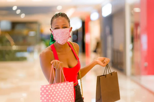 young African American woman at shopping mall in new normal after covid-19 - happy and beautiful black girl in face mask holding shopping bags enjoying at beauty fashion store