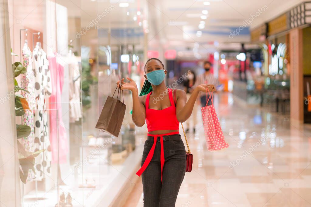 young African American woman at shopping mall in new normal after covid-19 - happy and beautiful black girl in face mask holding shopping bags enjoying at beauty fashion store 