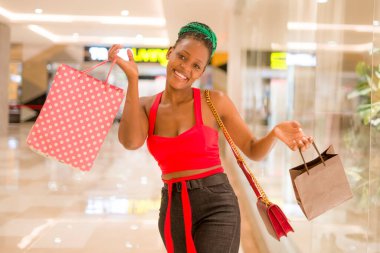 Genç, mutlu ve güzel siyah Afro-Amerikan kadının portresi alışveriş merkezinden alışveriş poşetleri alıyor, gülümsüyor ve şık kıyafetler içinde satışlardan keyif alıyor.