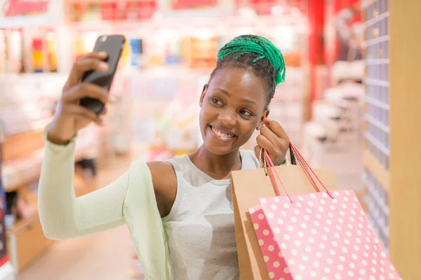 lifestyle portrait of young happy and beautiful black African American woman holding shopping bags taking selfie with mobile phone buying at mall fashion store smiling enjoying sales