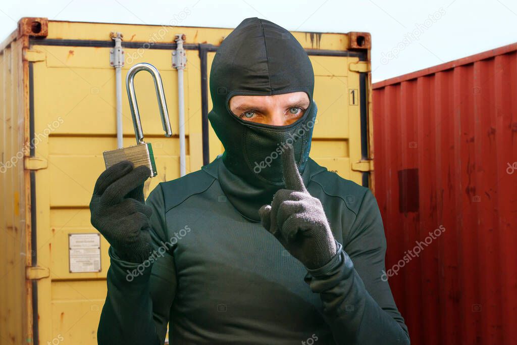 thief at work - criminal man in black covered with balaclava mask holding unlocked padlock at shipping area break in storage containers in robbery and crime concept