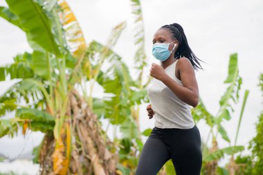 Karantina sonrası koşucu kız dışarıda antrenman yapıyor. Genç, çekici ve formda Afro-Amerikan bir kadın. Yeni normal spor konsepti ve sağlıklı yaşam tarzı ile yüz maskesi takıyor.