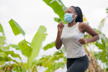 Karantina sonrası koşucu kız dışarıda antrenman yapıyor. Genç, çekici ve formda Afro-Amerikan bir kadın. Yeni normal spor konsepti ve sağlıklı yaşam tarzı ile yüz maskesi takıyor.