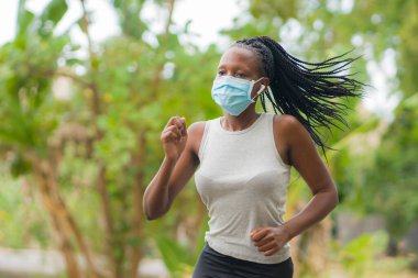 Karantina sonrası koşucu kız dışarıda antrenman yapıyor. Genç, çekici ve formda Afro-Amerikan bir kadın. Yeni normal spor konsepti ve sağlıklı yaşam tarzı ile yüz maskesi takıyor.