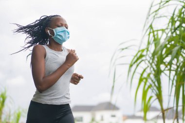 Karantina sonrası koşucu kız dışarıda antrenman yapıyor. Genç, çekici ve formda Afro-Amerikan bir kadın. Yeni normal spor konsepti ve sağlıklı yaşam tarzı ile yüz maskesi takıyor.