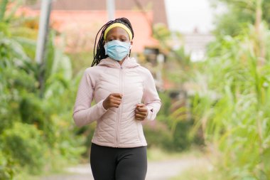 Karantina sonrası koşucu kız dışarıda antrenman yapıyor. Genç, çekici ve formda Afro-Amerikan bir kadın. Yeni normal spor konsepti ve sağlıklı yaşam tarzı ile yüz maskesi takıyor.