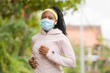 Karantina sonrası koşucu kız dışarıda antrenman yapıyor. Genç, çekici ve formda Afro-Amerikan bir kadın. Yeni normal spor konsepti ve sağlıklı yaşam tarzı ile yüz maskesi takıyor.