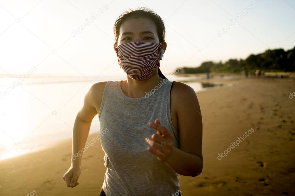 new normal running workout of Asian girl in face mask - young happy and attractive Chinese woman jogging on the beach in post quarantine outdoors exercise on beautiful sunset