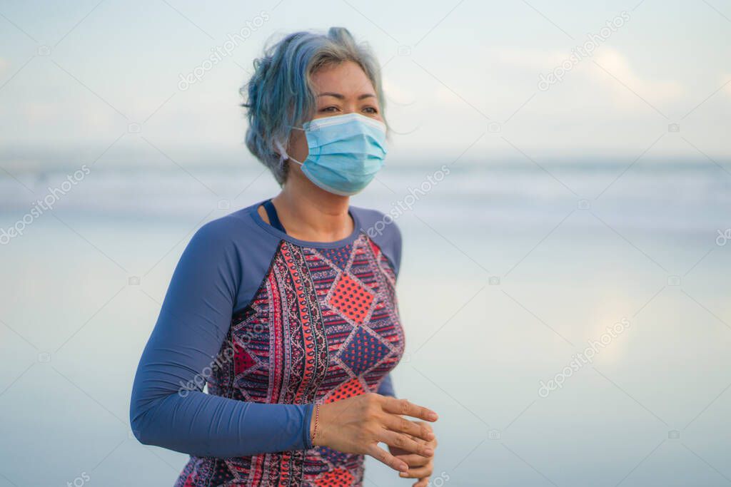 new normal running workout with face mask - attractive and happy middle aged woman on her 40s or 50s doing post quarantine jogging at beautiful beach in healthy lifestyle concept