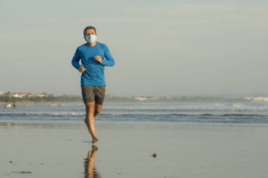 Yeni normal plaj koşusu antrenmanı. Genç fit ve çekici bir adam. Yüzü maske takarak denizde çıplak koşuyor. 