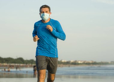 Yeni normal plaj koşusu antrenmanı. Genç fit ve çekici bir adam. Yüzü maske takarak denizde koşuyor.