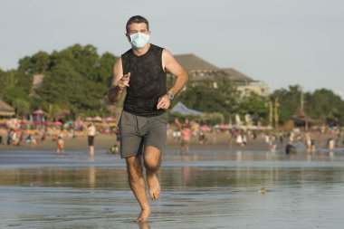 Yeni normal plaj koşusu egzersizi - sağlıklı yaşam tarzı konseptinde özgür hissederek deniz eğitiminden sonra çıplak ayak koşan yüzü maskeli genç ve çekici bir adam.