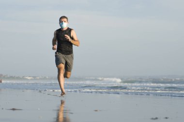 Yeni normal plaj koşusu egzersizi - sağlıklı yaşam tarzı konseptinde özgür hissederek deniz eğitiminden sonra çıplak ayak koşan yüzü maskeli genç ve çekici bir adam.