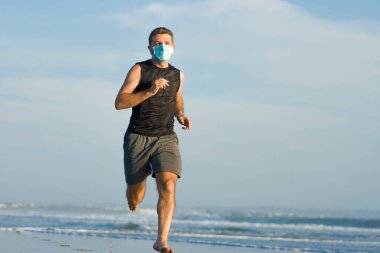 Yeni normal plaj koşusu egzersizi - sağlıklı yaşam tarzı konseptinde özgür hissederek deniz eğitiminden sonra çıplak ayak koşan yüzü maskeli genç ve çekici bir adam.