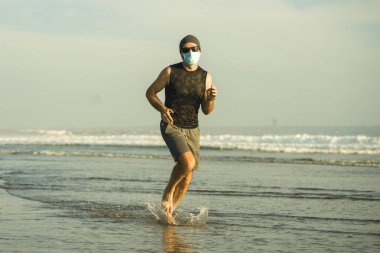 Yeni normal plaj koşusu egzersizi - sağlıklı yaşam tarzı konseptinde özgür hissederek deniz eğitiminden sonra çıplak ayak koşan yüzü maskeli genç ve çekici bir adam.