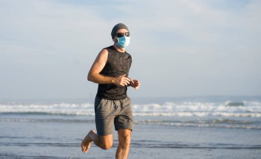 Yeni normal plaj koşusu egzersizi - sağlıklı yaşam tarzı konseptinde özgür hissederek deniz eğitiminden sonra çıplak ayak koşan yüzü maskeli genç ve çekici bir adam.