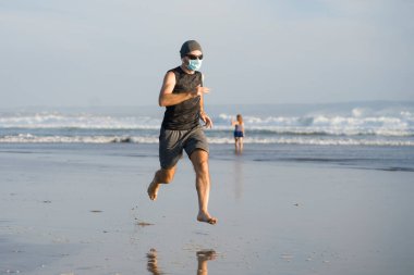 Yeni normal plaj koşusu egzersizi - sağlıklı yaşam tarzı konseptinde özgür hissederek deniz eğitiminden sonra çıplak ayak koşan yüzü maskeli genç ve çekici bir adam.