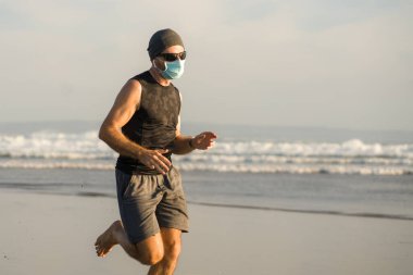 Yeni normal plaj koşusu egzersizi - sağlıklı yaşam tarzı konseptinde özgür hissederek deniz eğitiminden sonra çıplak ayak koşan yüzü maskeli genç ve çekici bir adam.