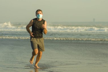 Yeni normal plaj koşusu egzersizi - sağlıklı yaşam tarzı konseptinde özgür hissederek deniz eğitiminden sonra çıplak ayak koşan yüzü maskeli genç ve çekici bir adam.