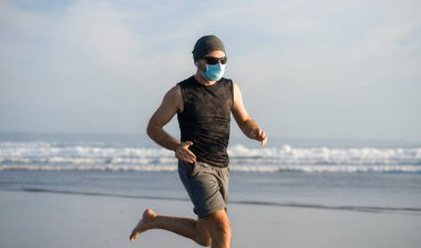 Yeni normal plaj koşusu egzersizi - sağlıklı yaşam tarzı konseptinde özgür hissederek deniz eğitiminden sonra çıplak ayak koşan yüzü maskeli genç ve çekici bir adam.