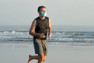 Yeni normal plaj koşusu egzersizi - sağlıklı yaşam tarzı konseptinde özgür hissederek deniz eğitiminden sonra çıplak ayak koşan yüzü maskeli genç ve çekici bir adam.