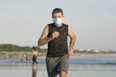 Yeni normal plaj koşusu egzersizi - sağlıklı yaşam tarzı konseptinde özgür hissederek deniz eğitiminden sonra çıplak ayak koşan yüzü maskeli genç ve çekici bir adam.