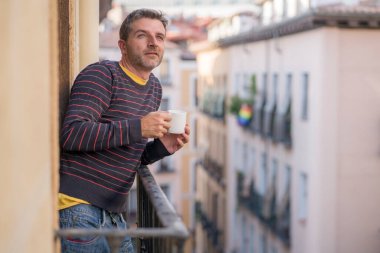 30 'lu ya da 40' lı yaşlarda çekici ve mutlu bir adam evde balkonda rahat ve neşeli bir fincan kahvenin keyfini çıkarıyor şehrin sokaklarına bakıp gülümsüyor ve şehir manzarasının tadını çıkarıyor.