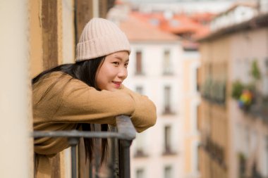 Avrupa 'da tatil gezisi sırasında İspanya' daki otel odasının balkonundan şehir manzarasının keyfini çıkaran, kışın şapkalı, mutlu ve güzel Asyalı bir kadın.