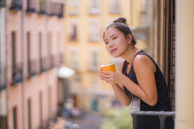 Genç, güzel ve mutlu Asyalı bir kadın İspanya 'da otel odası balkonundan şehir manzarasını seyrediyor Avrupa' da tatil gezisi sırasında kentsel arka planda rahatlamış kahve içiyor.