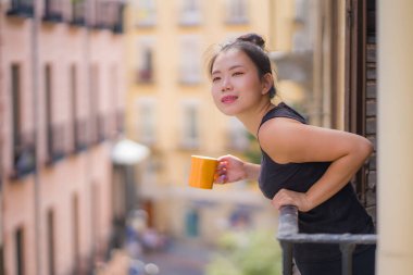 Genç, güzel ve mutlu Asyalı bir kadın İspanya 'da otel odası balkonundan şehir manzarasını seyrediyor Avrupa' da tatil gezisi sırasında kentsel arka planda rahatlamış kahve içiyor.