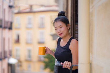 Genç, güzel ve mutlu Asyalı bir kadın İspanya 'da otel odası balkonundan şehir manzarasını seyrediyor Avrupa' da tatil gezisi sırasında kentsel arka planda rahatlamış kahve içiyor.