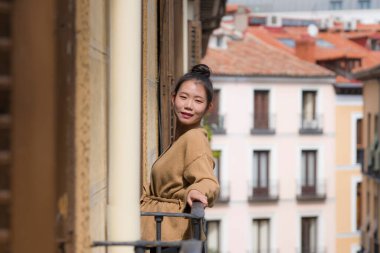Avrupa 'da tatil gezisi sırasında İspanya' da otel odası balkonundan şehir manzarasının keyfini çıkaran, saçları topuzlu, genç ve güzel Asyalı bir Çinli kadın kentsel arka planda neşeli bir şekilde gülümsüyor.