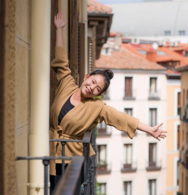 Avrupa 'da tatil gezisi sırasında İspanya' da otel odası balkonundan şehir manzarasının keyfini çıkaran, saçları topuzlu, genç ve güzel Asyalı bir Çinli kadın kentsel arka planda neşeli bir şekilde gülümsüyor.