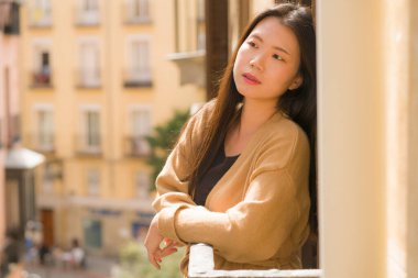 Genç mutlu ve güzel Asyalı Japon kadın İspanya 'da otel odasının balkonundan şehir manzarasının keyfini çıkarırken Avrupa' da tatil gezisi sırasında şehir arka planında neşeli bir şekilde gülümsüyor.