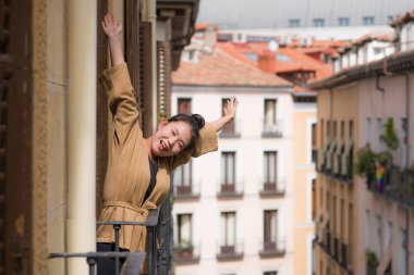 Avrupa 'da tatil gezisi sırasında İspanya' da otel odası balkonundan şehir manzarasının keyfini çıkaran, saçları topuzlu, genç ve güzel Asyalı bir Japon kadın kentsel arka planda neşeyle gülümsüyor.