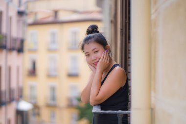 Avrupa 'da tatil gezisi sırasında İspanya' da otel odası balkonundan şehir manzarasının keyfini çıkaran, saçları topuzlu, genç ve güzel Asyalı bir Japon kadın kentsel arka planda neşeyle gülümsüyor.