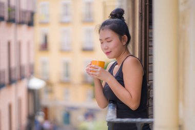 Genç, güzel ve mutlu Asyalı Çinli kadın İspanya 'da tatil gezisi sırasında otel odasının balkonundan şehir manzarasının keyfini çıkarıyor. Şehir arka planında rahatça kahve içiyor.