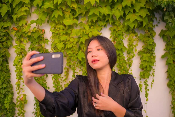 young happy and beautiful Asian Chinese woman enjoying outdoors taking selfie with mobile phone at village garden playful and carefree in beauty fashion and freedom concept