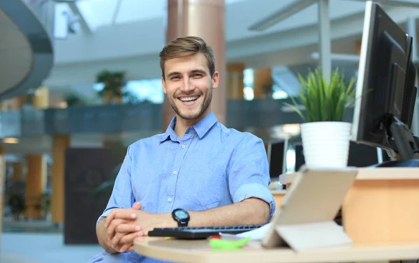 Portrait of happy man sitting at office desk, looking at camera, smiling.