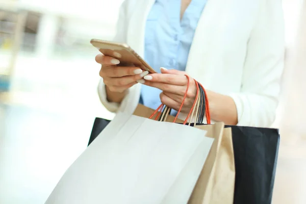 Mujer usando teléfono inteligente y sosteniendo la bolsa de compras mientras está de pie en el fondo del centro comercial . —  Fotos de Stock