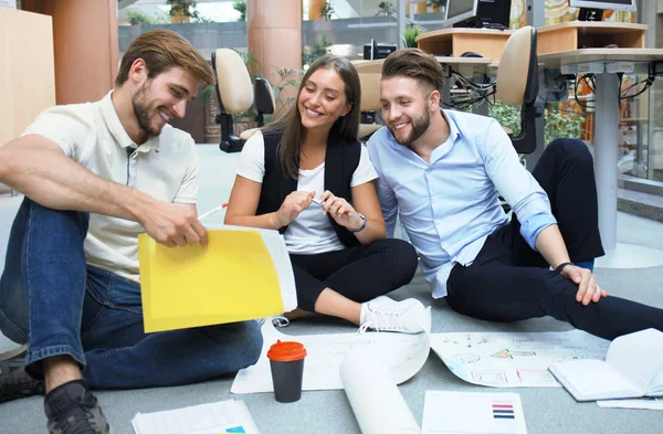 Grupo de jóvenes empresarios y diseñadores que miran el plan de proyecto dispuesto en el piso. Están trabajando en un nuevo proyecto . — Foto de Stock