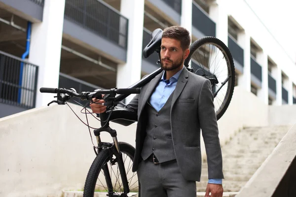 Jovem empresário confiante carregando sua bicicleta enquanto caminha ao ar livre . — Fotografia de Stock