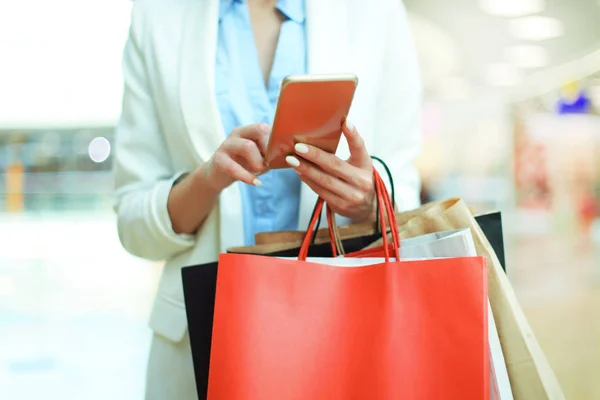 Vrouw met behulp van smartphone en boodschappentas terwijl staande te houden op de achtergrond winkelcentrum. — Stockfoto