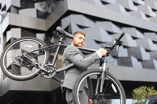 Jovem empresário confiante carregando sua bicicleta enquanto caminha ao ar livre . — Fotografia de Stock